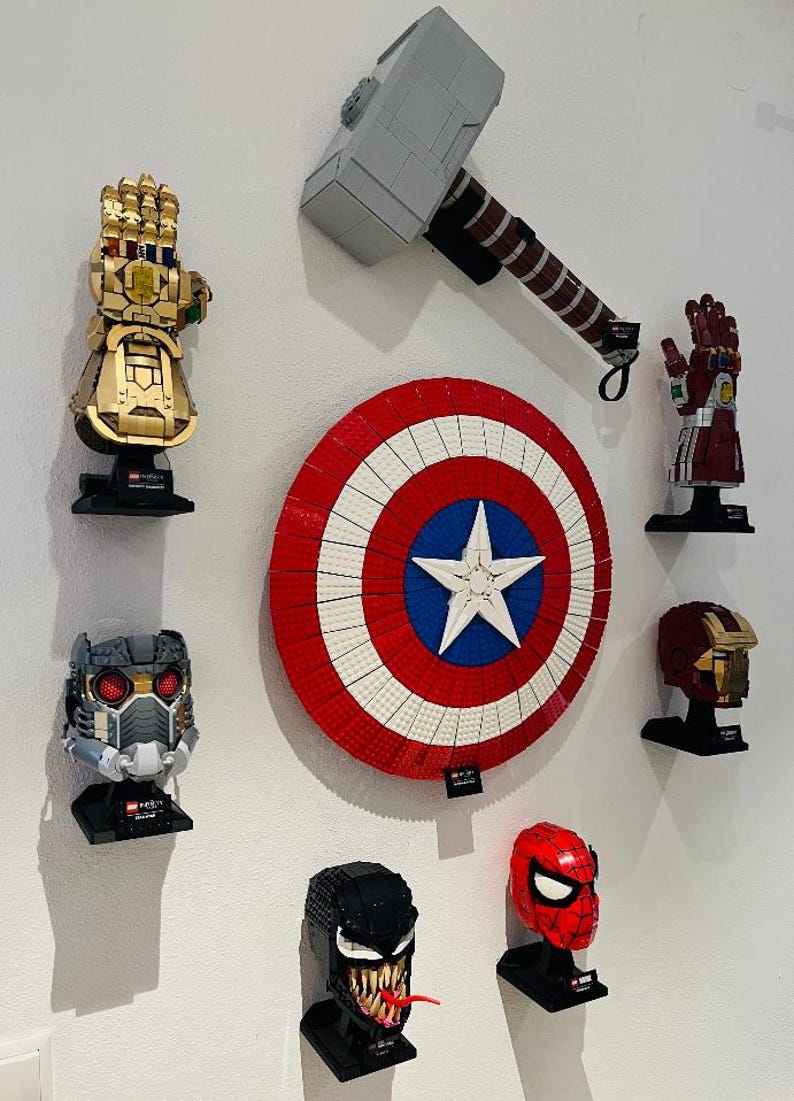 Puede incluir: Una colecci&oacute;n de construcciones Lego de personajes de Marvel montadas en una pared blanca. Incluye un Guantelete del Infinito dorado, el martillo de Thor, el escudo del Capit&aacute;n Am&eacute;rica, el casco de Iron Man y otros bustos de personajes.