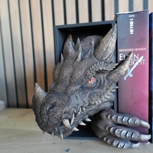 May include: A detailed dragon head bookend in shades of gray and brown. The dragon's head emerges from a black rectangular frame, with sharp teeth and red eyes. The bookend is designed to hold books, as seen with a book on the right side.