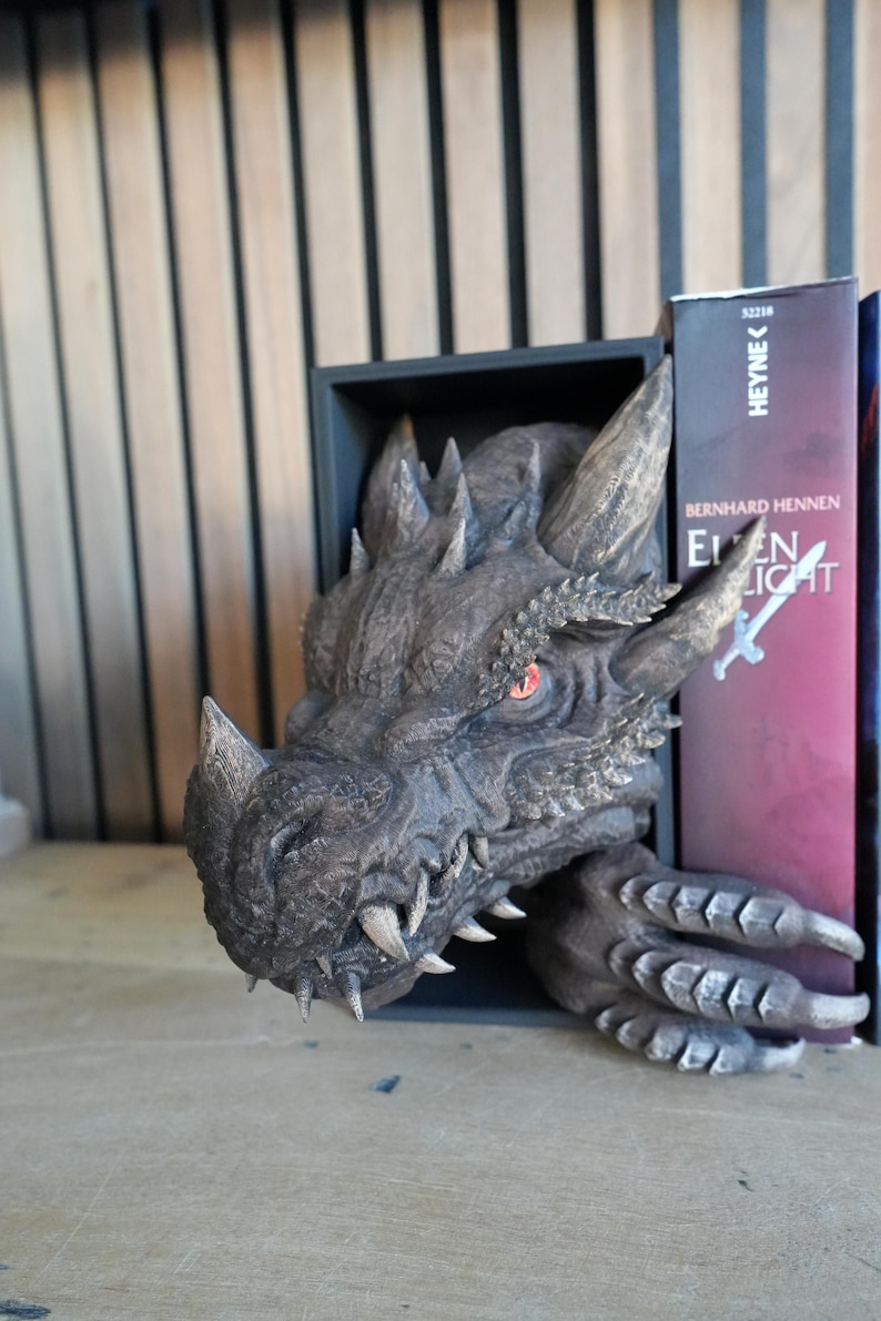 May include: A detailed dragon head bookend in shades of gray and brown. The dragon's head emerges from a black rectangular frame, with sharp teeth and red eyes. The bookend is designed to hold books, as seen with a book on the right side.