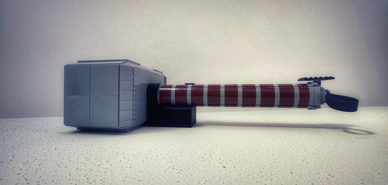 Op de afbeelding: Een grijze en bruine speelgoedhamer, die lijkt op Thor's Mjolnir, gemaakt van in elkaar grijpende blokken. De hamer heeft een rechthoekige kop en een lange, cilindrische steel. De steel is bruin met grijze strepen.