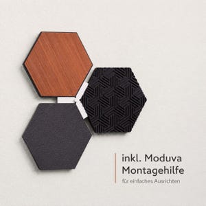 May include: Three hexagonal wall tiles in a modern design. One tile is wood-grained, another is textured black, and the third is a geometric black pattern. The tiles are connected with white connectors. Text in the image reads "inkl. Moduva Montagehilfe f&uuml;r einfaches Ausrichten."