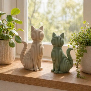 May include: Two decorative cat figurines. One is a light beige color, and the other is a muted green. Both are sitting upright with their tails curled. The cats have a ribbed design and are placed on a wooden surface.