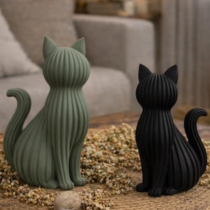May include: Two decorative cat figurines. One is sage green, the other is black. Both are seated with upright ears and curved tails. The cats have a ribbed texture and are placed on a woven mat with dried flowers.