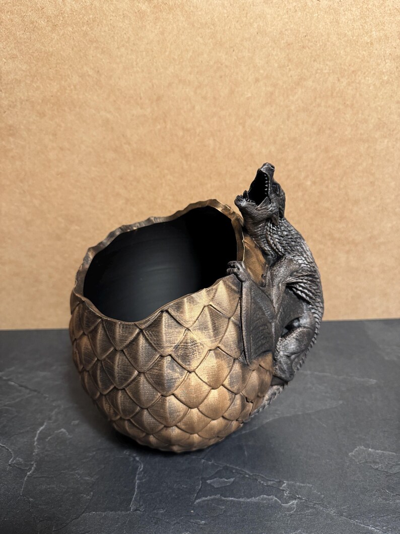 May include: A bronze-colored dragon egg-shaped planter with a black interior. A detailed dragon figure is perched on the side, its mouth open. The egg has a textured, scale-like pattern. The planter is on a dark surface.