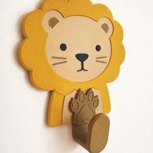May include: A wall-mounted lion-shaped hook featuring a yellow mane, a beige face with black eyes and nose, and a brown paw-shaped base. The hook itself is a brown, curved, upward-facing structure extending from the bottom of the lion figure.