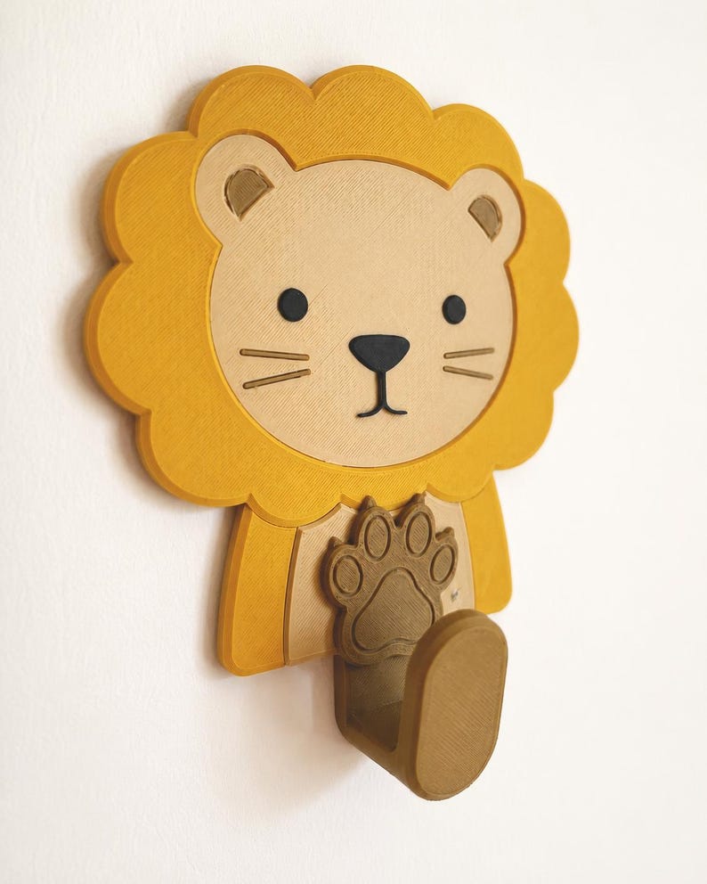 May include: A wall-mounted lion-shaped hook featuring a yellow mane, a beige face with black eyes and nose, and a brown paw-shaped base. The hook itself is a brown, curved, upward-facing structure extending from the bottom of the lion figure.