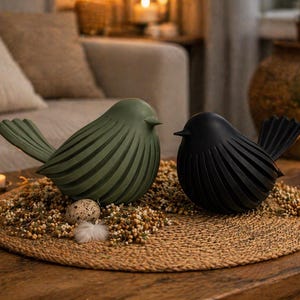 May include: Two decorative bird sculptures, one olive green and one black, sit on a round woven mat. The birds have a stylized design with ribbed detailing. The mat is adorned with small, round, beige decorative elements and a small egg.