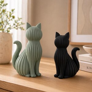 May include: Two decorative cat figurines. One is sage green, the other is black. Both are sitting upright with their tails curled. The cats have a ribbed texture and are placed on a wooden surface.