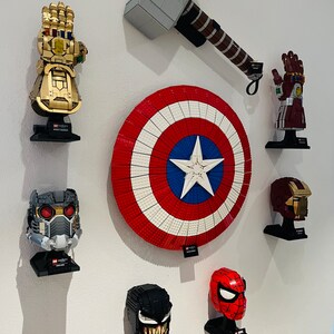 May include: A collection of Marvel-themed LEGO sets mounted on a white wall. The display includes a gold Infinity Gauntlet, Captain America's shield, Thor's hammer, and various character helmets, all on black stands.