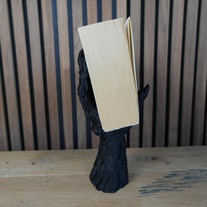 May include: A black, stylized book holder in the shape of a tree trunk with outstretched branches supporting a book. The book has light-colored pages and a beige cover. The holder is on a wooden surface.