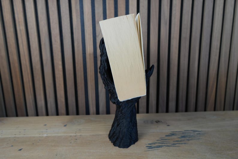 May include: A black, stylized book holder in the shape of a tree trunk with outstretched branches supporting a book. The book has light-colored pages and a beige cover. The holder is on a wooden surface.
