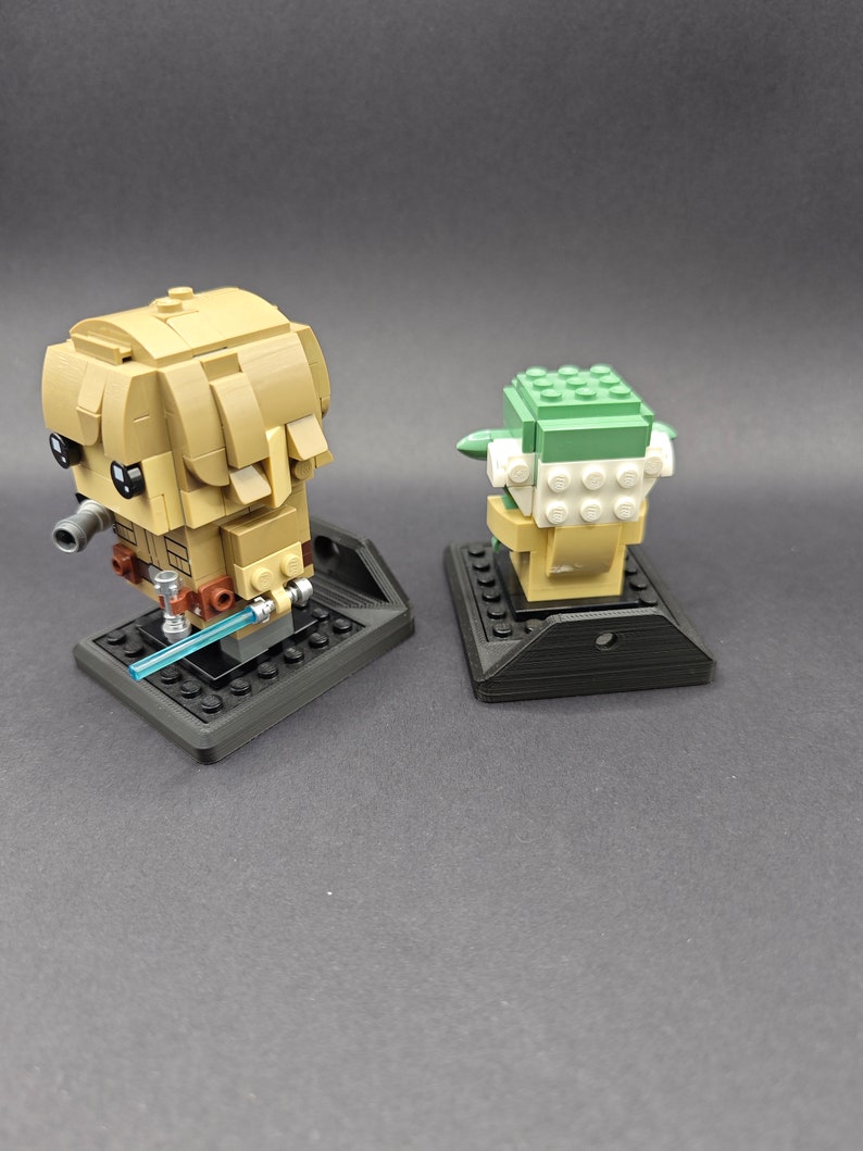 Może przedstawiać: Dwie minifigurki Lego, jedna przedstawiająca Yodę, a druga Jedi, są wystawione na czarnych podstawkach Lego. Figurki są zwr&oacute;cone do siebie.