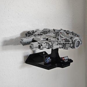 以下が含まれることがあります： 黒い壁掛けディスプレイスタンドに設置された、グレーとブラックのLEGOミレニアム・ファルコンのスターシップモデル。スタンドにはスターウォーズのロゴと25周年記念のロゴが描かれています。