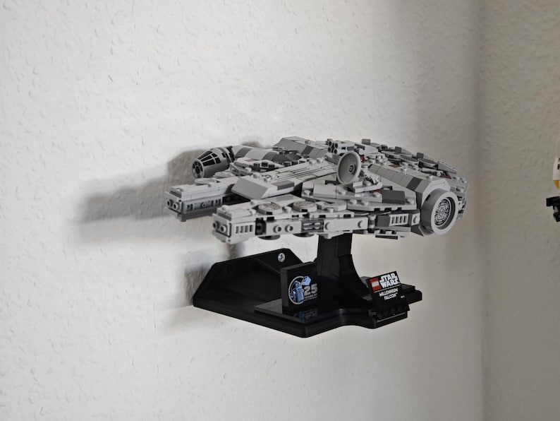 以下が含まれることがあります： 黒い壁掛けディスプレイスタンドに設置された、グレーとブラックのLEGOミレニアム・ファルコンのスターシップモデル。スタンドにはスターウォーズのロゴと25周年記念のロゴが描かれています。