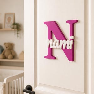 May include: A large, magenta-colored wooden letter N mounted on a white door, featuring the name nami in white, lowercase, three-dimensional script lettering layered directly over the center of the letter N.