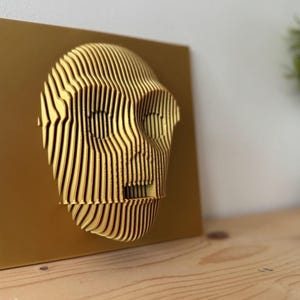 Puede incluir: Una escultura abstracta de rostro en 3D impresa en oro, montada sobre un fondo dorado.