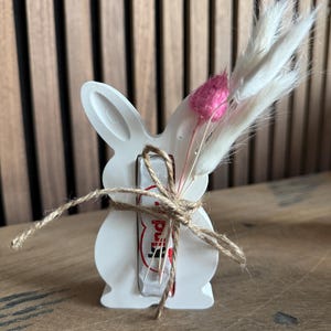 May include: White ceramic bunny figurine holding a chocolate bar wrapped in red and white foil. The bunny is decorated with a pink and white dried flower arrangement and twine.