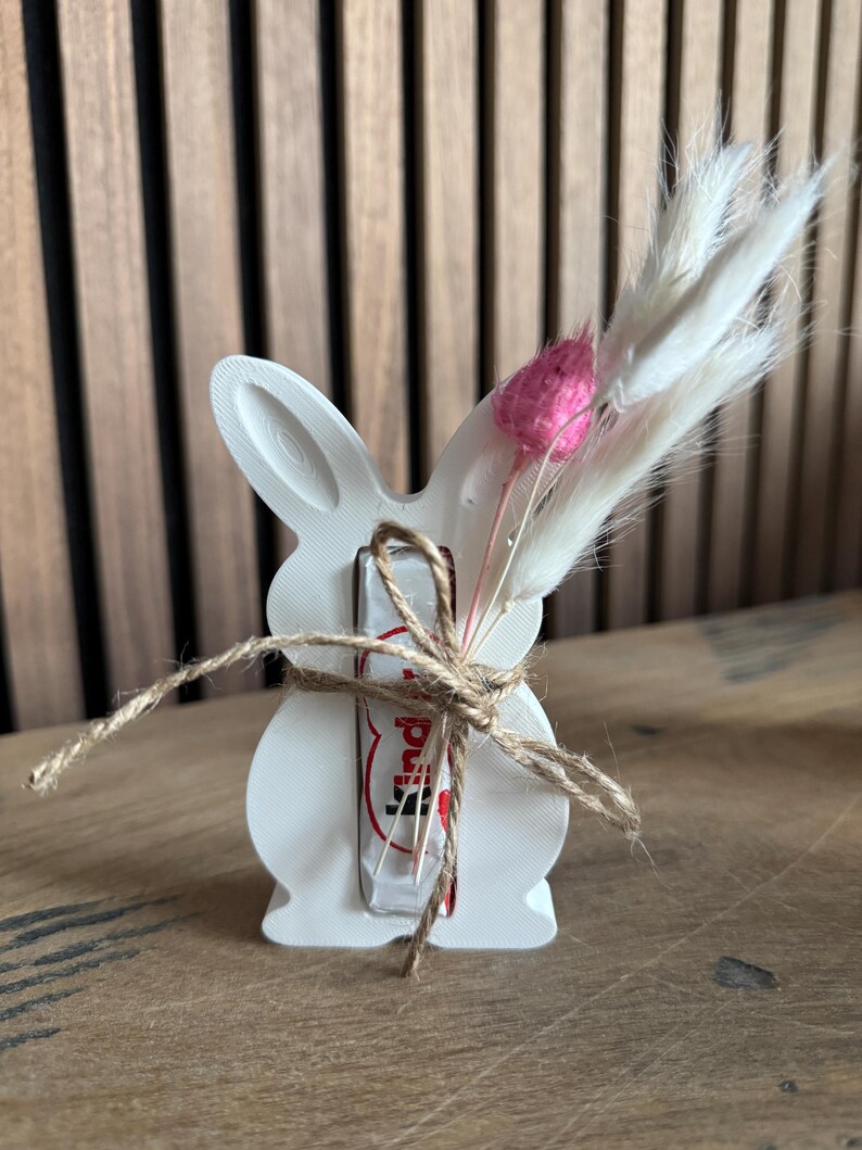 May include: White ceramic bunny figurine holding a chocolate bar wrapped in red and white foil. The bunny is decorated with a pink and white dried flower arrangement and twine.