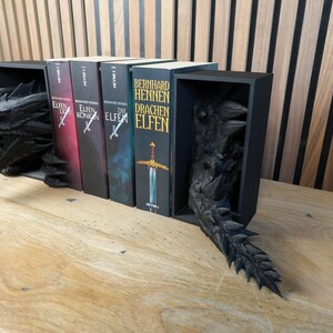 May include: Black dragon bookends with a detailed, textured design. The bookends hold several books with titles in German. The dragon's head and tail emerge from the black bookends, creating a fantasy theme.