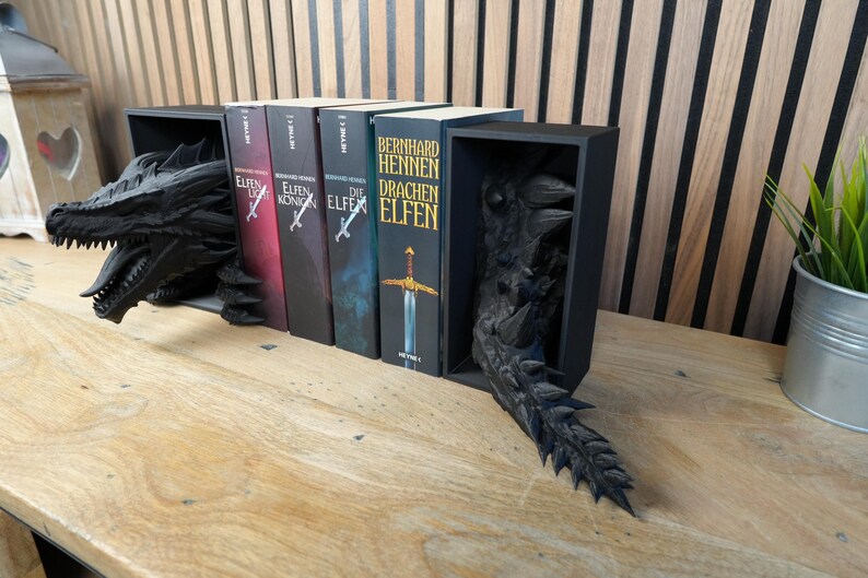 May include: Black dragon bookends with a detailed, textured design. The bookends hold several books with titles in German. The dragon's head and tail emerge from the black bookends, creating a fantasy theme.