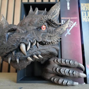 May include: A detailed dragon head bookend in shades of gray and brown, with sharp teeth and a red eye. The dragon's claw rests on a shelf next to several books. A unique decorative item for fantasy book lovers.