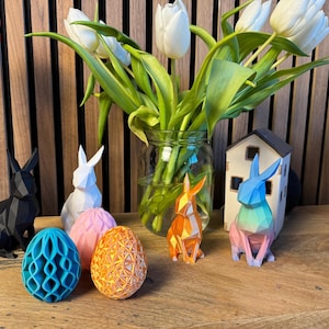 May include: Five origami paper animals, including a black rabbit, a white rabbit, a blue rabbit, an orange rabbit, and a teal egg, are displayed on a wooden surface. A vase of white tulips is in the background.
