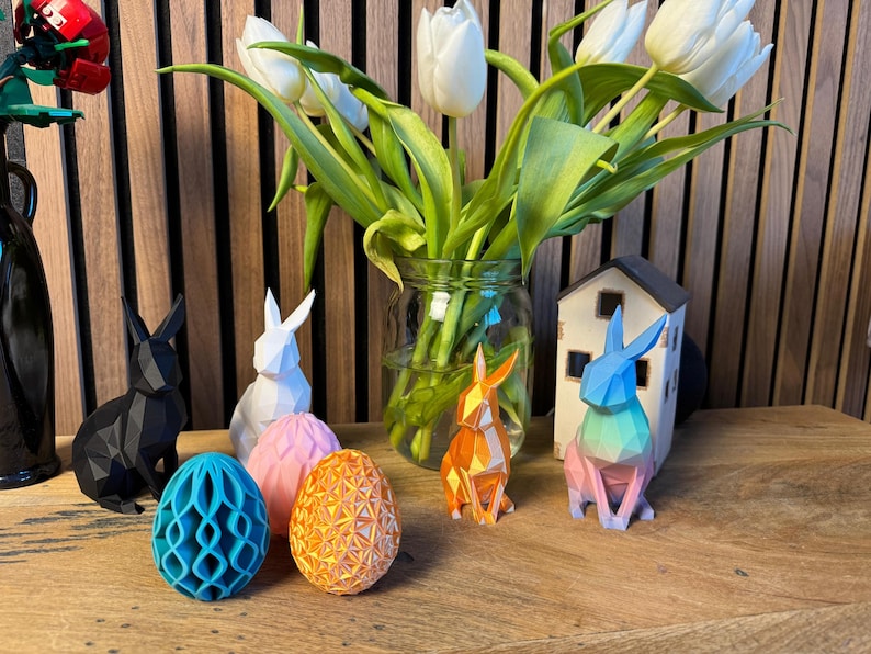 May include: Five origami paper animals, including a black rabbit, a white rabbit, a blue rabbit, an orange rabbit, and a teal egg, are displayed on a wooden surface. A vase of white tulips is in the background.