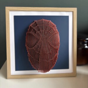 May include: A framed wall art piece featuring a 3D abstract sculpture made of thin, vertical, red lines. The sculpture is circular and appears to be made of wood. The sculpture is mounted on a dark blue background.