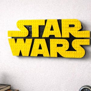 Pode incluir: Logotipo amarelo "STAR WARS" feito de blocos de brinquedo, montado em uma parede branca. O logotipo est&aacute; em uma fonte arrojada e em blocos. Uma pequena planta em vaso e material de escrit&oacute;rio s&atilde;o vis&iacute;veis abaixo.