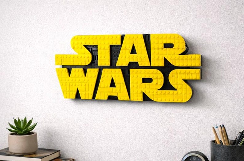 Pode incluir: Logotipo amarelo "STAR WARS" feito de blocos de brinquedo, montado em uma parede branca. O logotipo est&aacute; em uma fonte arrojada e em blocos. Uma pequena planta em vaso e material de escrit&oacute;rio s&atilde;o vis&iacute;veis abaixo.