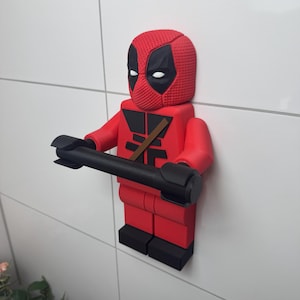 May include: A red and black Deadpool Lego figurine holding a black toilet paper roll holder. The figurine is mounted on a white tiled wall.