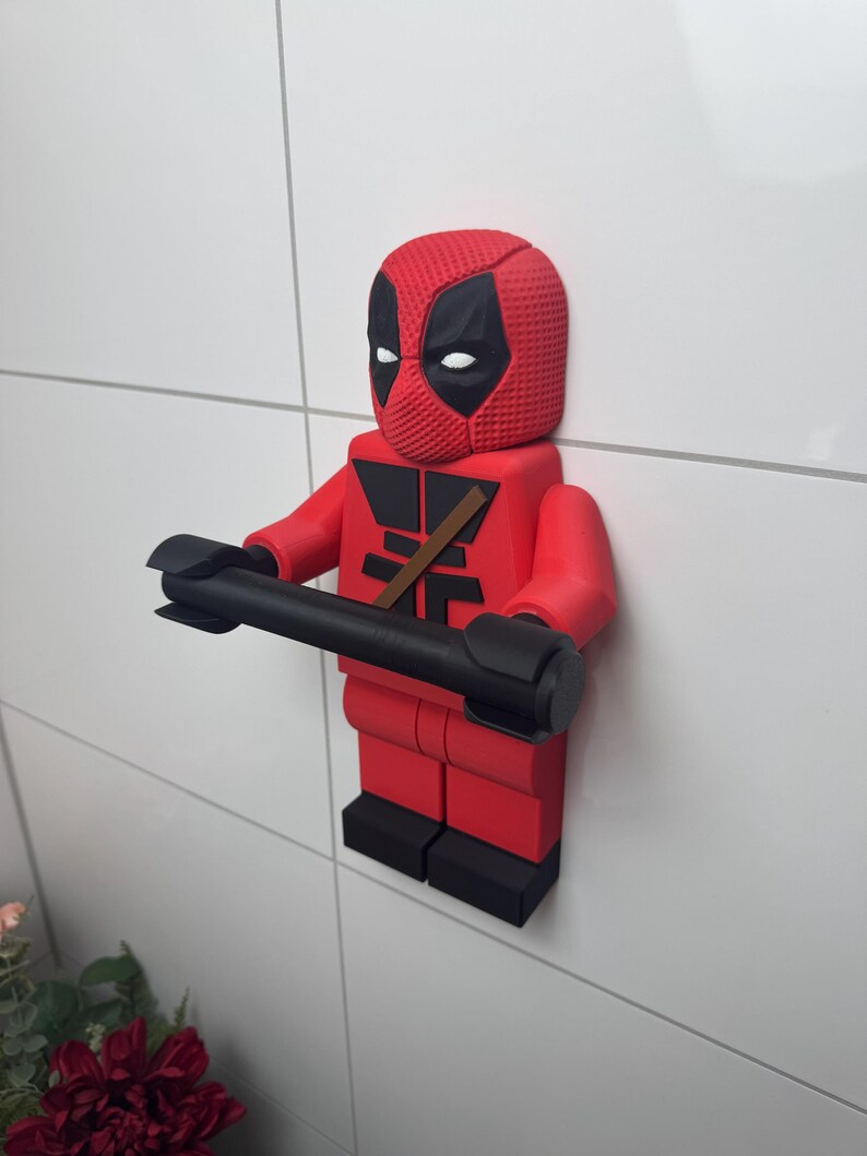 May include: A red and black Deadpool Lego figurine holding a black toilet paper roll holder. The figurine is mounted on a white tiled wall.