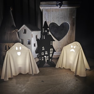 Puede incluir: Dos portavelas blancos en forma de fantasma con caras felices y tristes, una silueta negra de una casa encantada y una caja de madera con un recorte en forma de coraz&oacute;n.