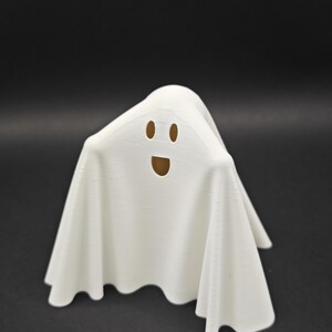 Puede incluir: Figurita de fantasma blanco con una cara sonriente. El fantasma est&aacute; hecho de un material blanco liso y tiene una forma fluida y drapeada.