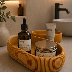 May include: A mustard yellow, ribbed, oval-shaped tray holds a brown Aesop bottle, a jar of Meraki body butter, and a tube of Meraki hand cream. The Aesop bottle has a dropper. The Meraki jar and tube have white labels.