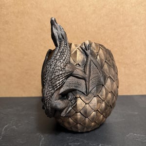 May include: A bronze-colored dragon figurine emerging from a textured egg. The dragon has detailed scales and wings, with its head and front legs visible. The egg has a scale-like pattern.