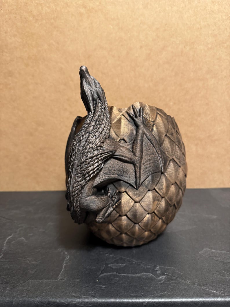 May include: A bronze-colored dragon figurine emerging from a textured egg. The dragon has detailed scales and wings, with its head and front legs visible. The egg has a scale-like pattern.