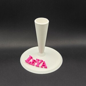 Puede incluir: Soporte para pastel blanco con un centro en forma de cono y una base redonda. La base tiene el nombre "LEYA" escrito en letras rosas.