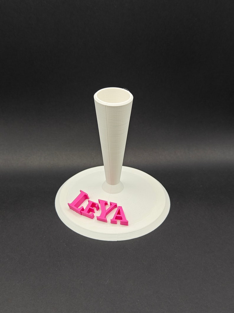 Puede incluir: Soporte para pastel blanco con un centro en forma de cono y una base redonda. La base tiene el nombre "LEYA" escrito en letras rosas.