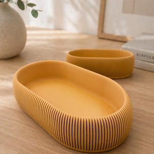 May include: Two mustard-yellow, oval-shaped trays. The larger tray is in the foreground, with a ribbed exterior and smooth interior. The smaller tray is in the background. Both trays are on a light-colored wooden surface.