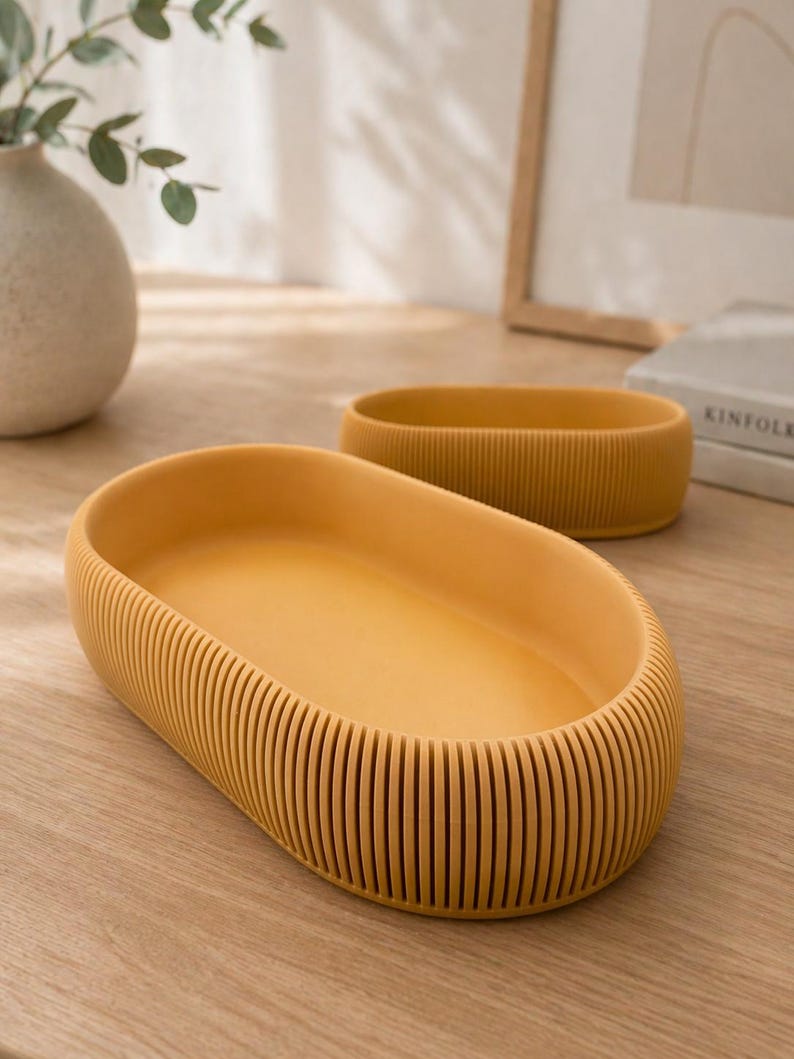 May include: Two mustard-yellow, oval-shaped trays. The larger tray is in the foreground, with a ribbed exterior and smooth interior. The smaller tray is in the background. Both trays are on a light-colored wooden surface.