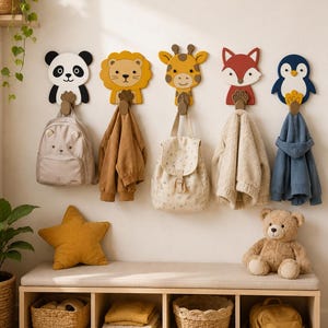 May include: Five wall-mounted hooks shaped like animal faces including a panda, lion, giraffe, fox, and penguin. Each hook features a brown paw-shaped base holding items such as a beige backpack, a brown jacket, a patterned backpack, a cream fleece jacket, and a blue hoodie.