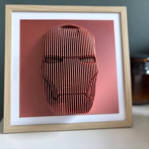 May include: A framed 3D wall art piece featuring a red, stylized face with a geometric design. The face is made of thin, vertical lines and is set against a red background.