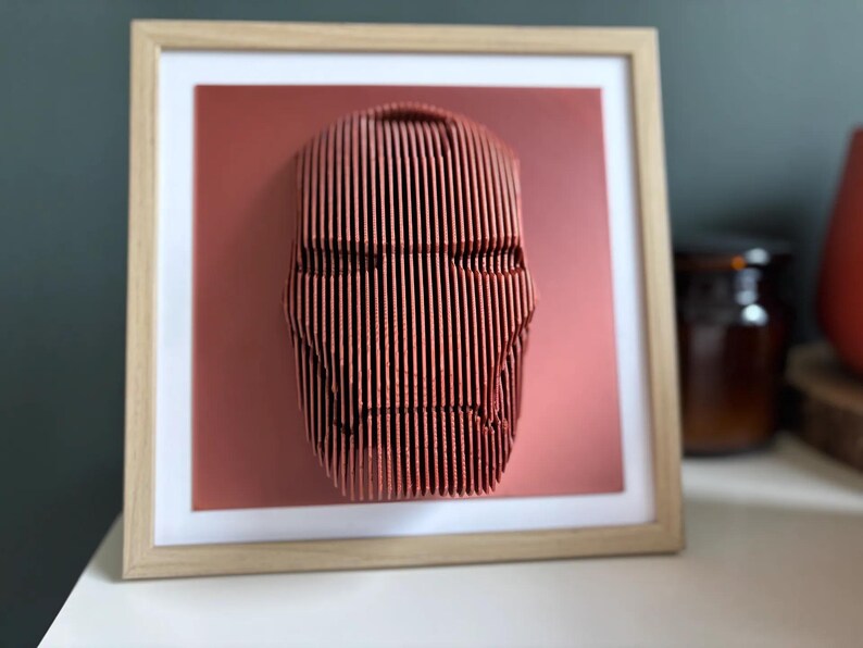 May include: A framed 3D wall art piece featuring a red, stylized face with a geometric design. The face is made of thin, vertical lines and is set against a red background.