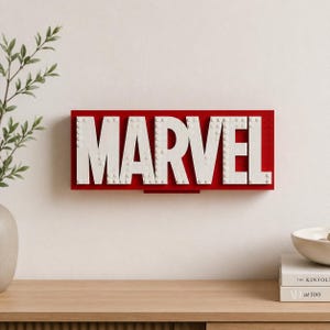 May include: A red rectangular sign with the word "MARVEL" in large white letters. The letters appear to be constructed from white blocks, and the sign is mounted on a white wall. The sign is a decorative item.