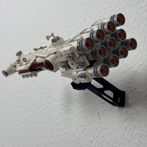 May include: A detailed model spaceship made from white and grey building blocks, with red detailing and multiple engine nozzles with orange centres. The model is mounted on a black stand against a white wall.