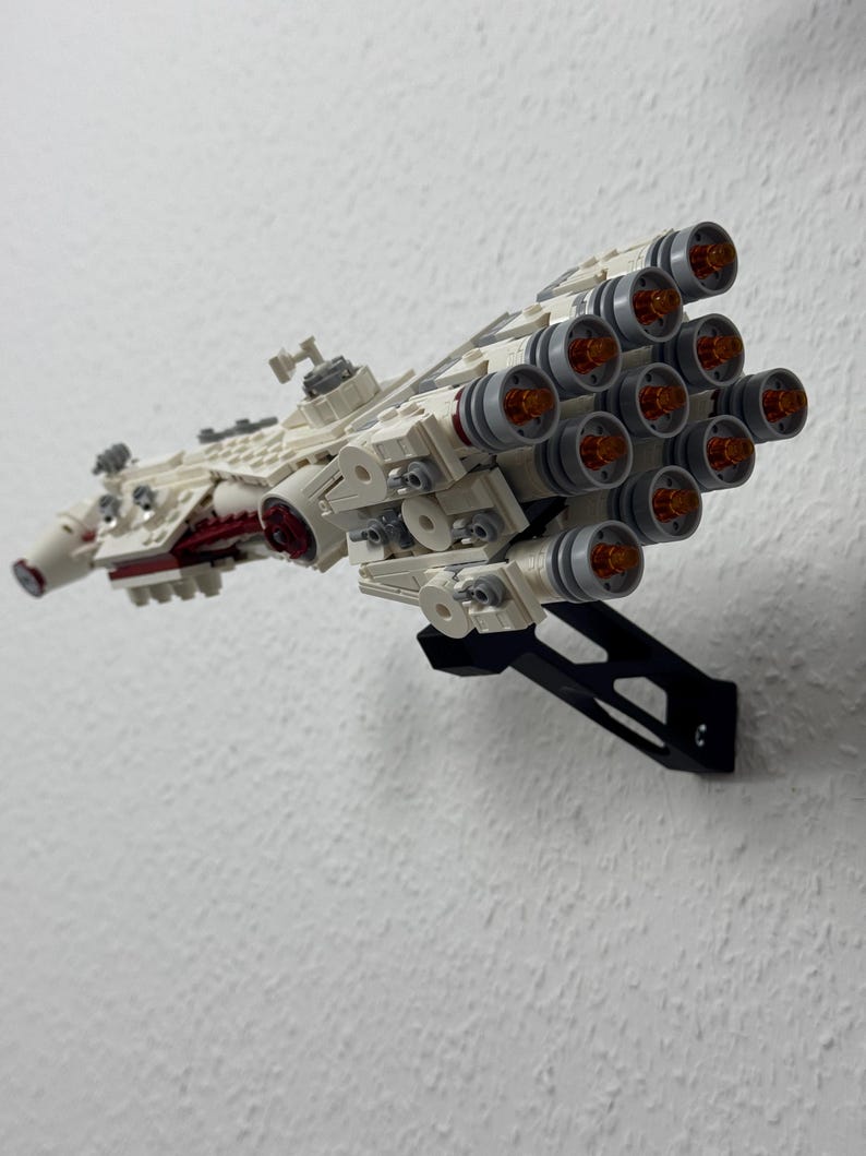 May include: A detailed model spaceship made from white and grey building blocks, with red detailing and multiple engine nozzles with orange centres. The model is mounted on a black stand against a white wall.
