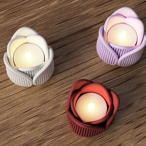 May include: Three decorative candle holders shaped like roses, each with a lit candle inside. One is white, one is purple, and the other is red. The holders are on a wooden surface.