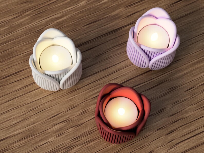 May include: Three decorative candle holders shaped like roses, each with a lit candle inside. One is white, one is purple, and the other is red. The holders are on a wooden surface.