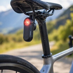Może przedstawiać: Czarna lampka tylna do roweru Garmin jest zamontowana pod czarnym siodełkiem rowerowym. Światło jest prostokątne z czerwoną soczewką i logo Garmin. Rower jest szary z czarną sztycą i oponą.